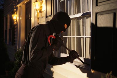 A masked burgler in the cloak of darkness using a crowbar on a window