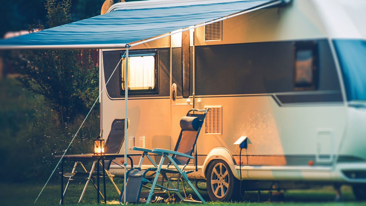 RV parked in a camp spot at evening time awning extened with two chairs, and a table with lantern on top.