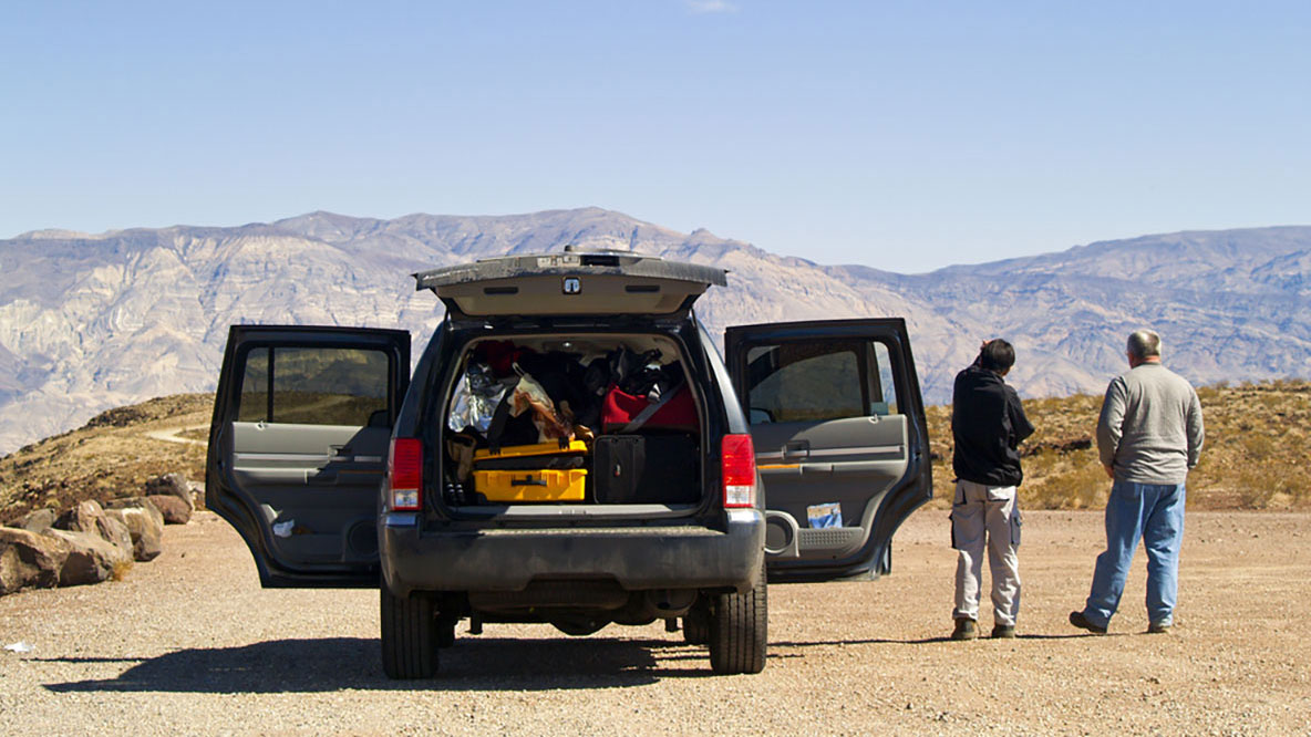 A suitcase filled suv stopped on a rural road with doors and back gate open. Two men standing outside of the car admiring the airid mountains in the distance.